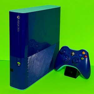 Könnte beinhalten: Eine blaue Xbox 360-Konsole und ein passender Controller werden vor einem leuchtend grünen Hintergrund gezeigt. Die Konsole hat das Xbox 360-Logo und ein Laufwerk. Der Controller steht auf einem schwarzen Ständer.