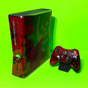 Puede incluir: Una consola Xbox 360 roja y verde con un mando a juego. La consola y el mando presentan un diseño de Gears of War con gráficos de calaveras. El mando descansa sobre un soporte negro.