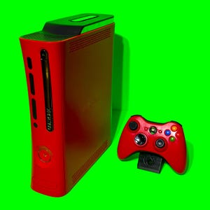 Puede incluir: Una consola Xbox 360 roja y un mando a juego. La consola tiene una parte superior negra y un cuerpo rojo. El mando tiene varios botones de colores y una base negra.