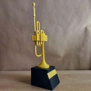 Peut inclure: Un trophée en forme de trompette de couleur dorée sur un socle noir avec une plaque de nom dorée. La trompette est stylisée et a un design simple.