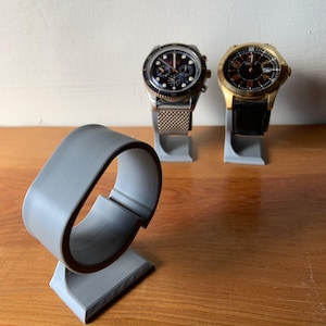 May include: Three gray watch stands for displaying wristwatches. Two stands have watches on them, one with a silver band and a black face, the other with a black band and a gold face.