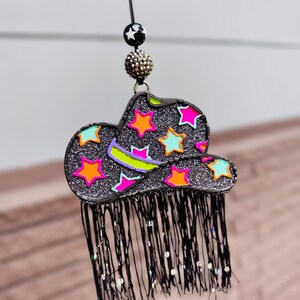 May include: A black glittery cowboy hat ornament featuring neon pink, orange, and light blue star accents. The hat has a black fringe trim hanging from the brim and is suspended by a black cord with a black and white star bead and a textured bead.