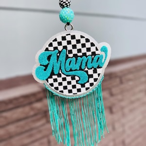 May include: A circular hanging ornament featuring a black and white checkered pattern with the word Mama in glittery teal script. The piece includes a teal fringe, a teal glitter bead, and a black and white checkered bead on a black cord.
