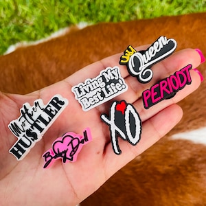 May include: A set of six colorful shoe charms with various phrases and designs, including "Mother Hustler", "Living My Best Life", "Queen", "Period", "Baby Doll", and "XO".