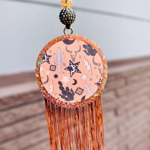 May include: A circular hanging ornament featuring a peach-colored background with black and white patterns of cacti, cow skulls, stars, lightning bolts, and geometric shapes. The edge is glittery orange, and it has a long, copper-colored fringe.