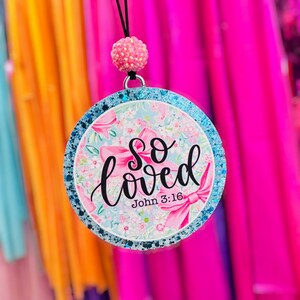 May include: A circular hanging ornament featuring a light blue background with pink bows and floral patterns. The text "so loved" is written in black script, with "John 3:16" below it. The border is blue glitter, topped with a pink textured bead.