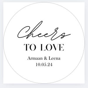 K&ouml;nnte beinhalten: Ein wei&szlig;es, kreisf&ouml;rmiges Design mit den Worten "Cheers TO LOVE" in schwarzer Schrift und Blockbuchstaben. Darunter stehen die Namen "Armaan & Leena" und das Datum "10.05.24" in einer einfachen Schriftart.