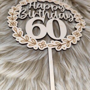 May include: Wooden cake topper with a floral wreath design. The topper says "Happy Birthday 60" in a cursive font.