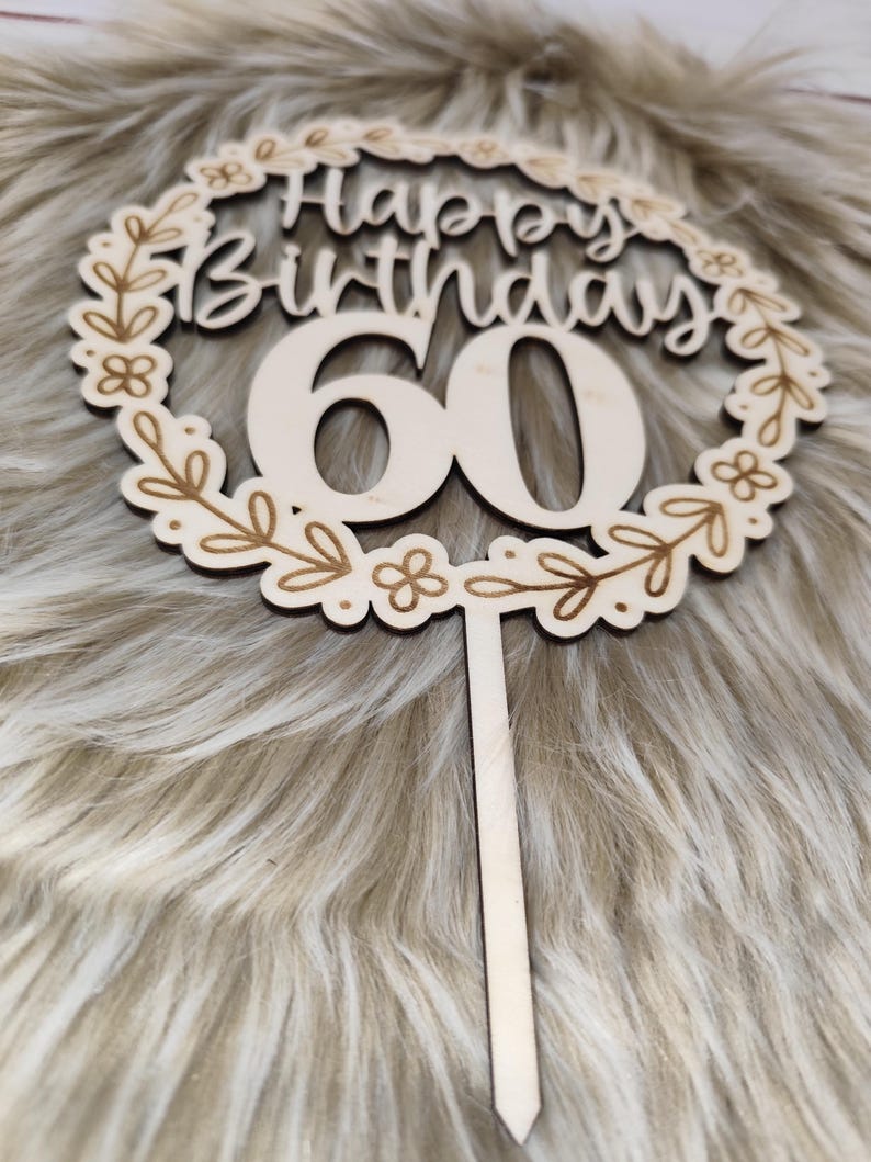 May include: Wooden cake topper with a floral wreath design. The topper says "Happy Birthday 60" in a cursive font.