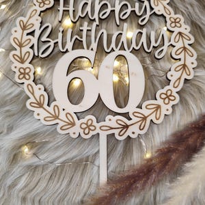 May include: Wooden cake topper with a floral wreath design. The topper says "Happy Birthday 60" in a white script font.