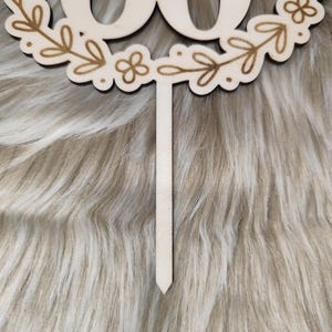 May include: Wooden cake topper with the number 60 in a wreath of leaves and flowers. The topper is on a white fuzzy background.