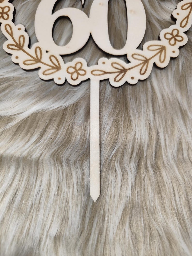 May include: Wooden cake topper with the number 60 in a wreath of leaves and flowers. The topper is on a white fuzzy background.