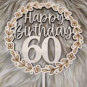 May include: Wooden cake topper with a floral wreath design. The topper says "Happy Birthday 60" in a cursive font.