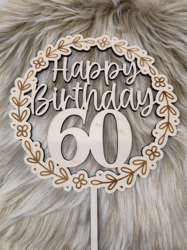 May include: Wooden cake topper with a floral wreath design. The topper says "Happy Birthday 60" in a cursive font.
