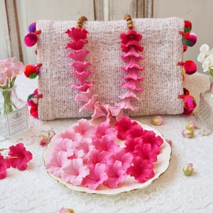 Puede incluir: Un clutch decorativo con detalles florales rosas y rojos, pompones y un plato de flores rosas. El clutch está hecho de una tela tejida. Pequeños jarrones con flores a ambos lados.