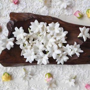 Puede incluir: Una colección de pequeñas flores de jazmín blancas de múltiples pétalos dispuestas dentro de un cuenco de madera de color marrón oscuro con bordes naturales. Las flores tienen una forma estrellada con pétalos delicados y superpuestos.