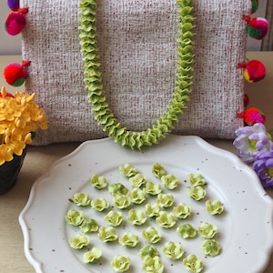 Puede incluir: Un collar de flores lei de color verde claro sobre un bolso de tela beige texturizada con pompones coloridos en los laterales, acompañado de un plato blanco festoneado con numerosas cuentas pequeñas individuales en forma de flor.