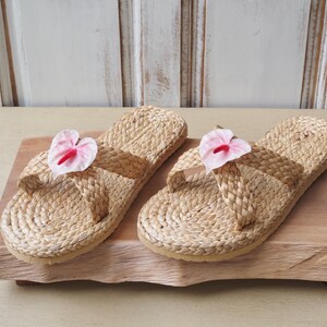 May include: Two pairs of woven straw sandals with pink flower embellishments. The sandals are on a wooden surface.