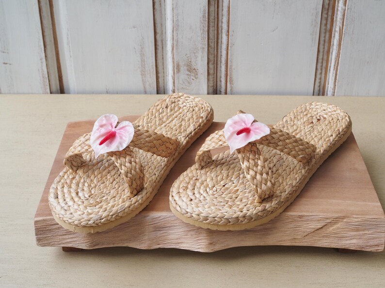May include: Two pairs of woven straw sandals with pink flower embellishments. The sandals are on a wooden surface.