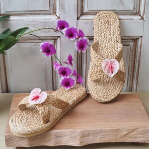 May include: Two pairs of woven straw sandals with pink flower accents. The sandals are on a wooden platform.