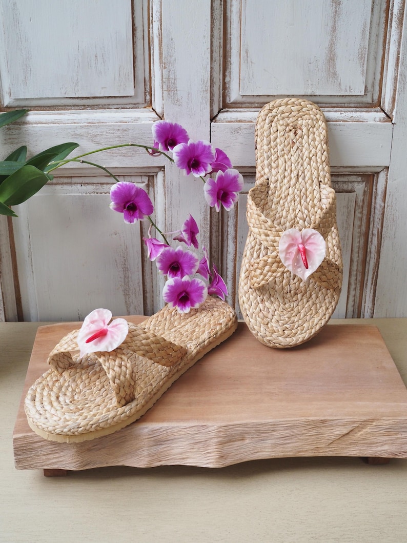 May include: Two pairs of woven straw sandals with pink flower accents. The sandals are on a wooden platform.