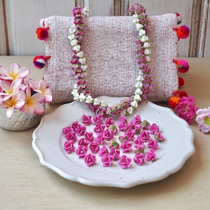 May include: A floral lei with white and purple flowers, draped over a woven cushion with pom-pom detailing. A white plate holds small pink rosebuds. A bowl of pink and yellow flowers is nearby.