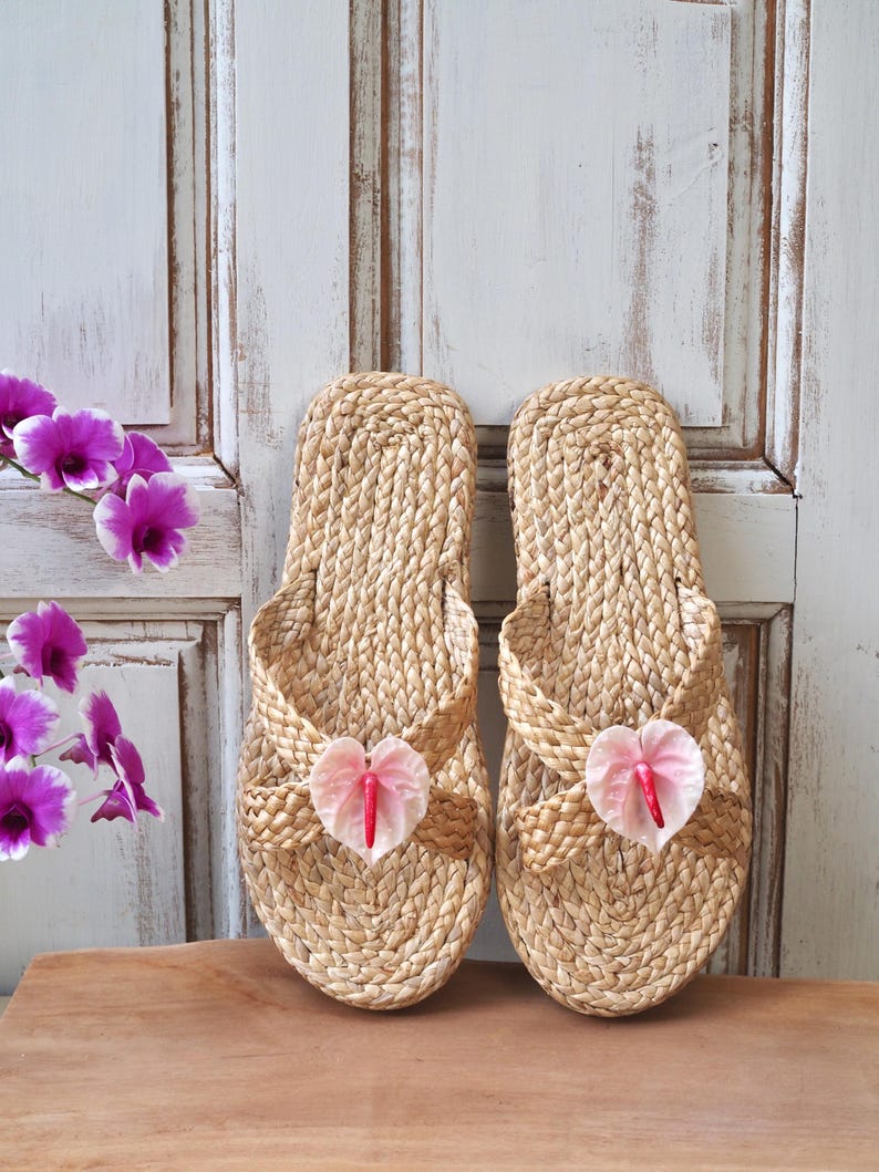 May include: A pair of woven straw sandals with pink flower accents. The sandals have a simple design and are perfect for warm weather.