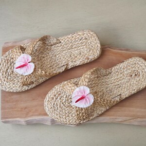 May include: Two pairs of woven straw sandals with pink flower embellishments. The sandals are on a wooden surface.