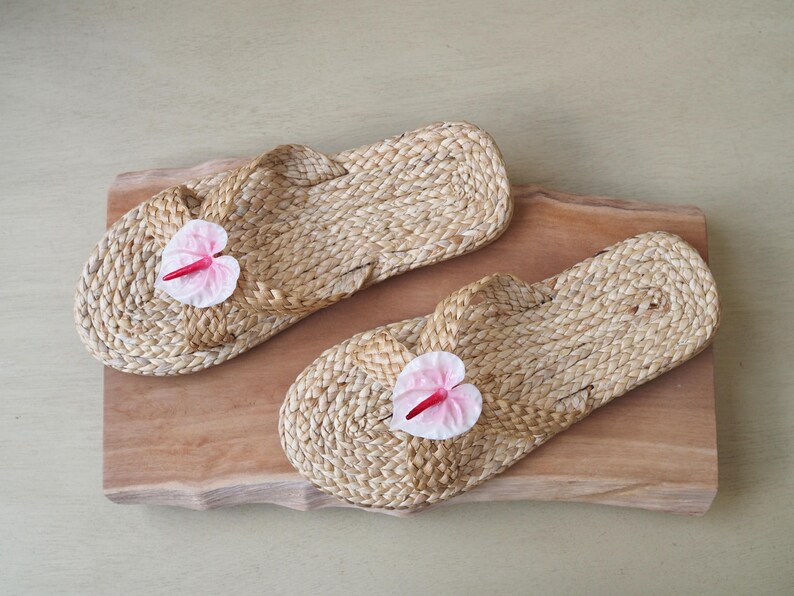 May include: Two pairs of woven straw sandals with pink flower embellishments. The sandals are on a wooden surface.