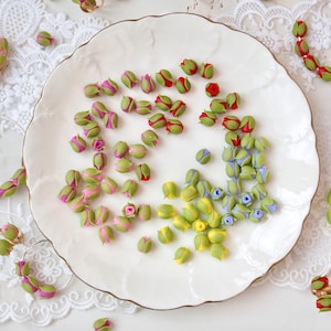 Op de afbeelding: Assortiment van kleine, handgemaakte polymer clay rozenknoppen in verschillende kleuren, waaronder roze, rood, geel, blauw en groen. De knoppen zijn gerangschikt op een wit bord.