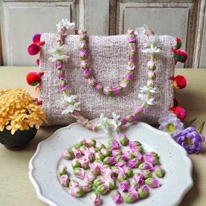 Puede incluir: Un bolso de tela rosa claro texturizada con pompones coloridos en los laterales, adornado con una larga guirnalda de capullos de rosa y flores de jazmín blancas. Un plato blanco festoneado contiene una colección de cuentas de capullos de rosa.