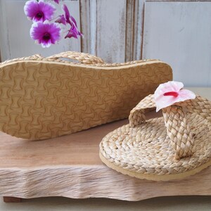 May include: Two pairs of woven straw sandals with a pink flower accent on one sandal. The sandals are on a wooden surface.