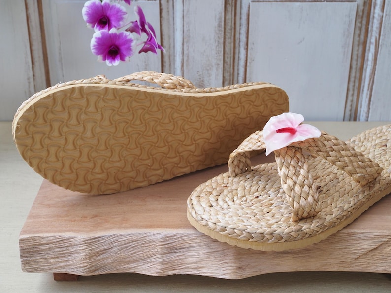 May include: Two pairs of woven straw sandals with a pink flower accent on one sandal. The sandals are on a wooden surface.