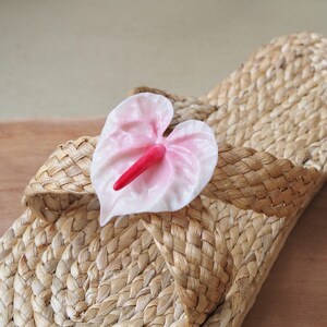May include: A pair of woven straw sandals with a pink flower accent. The flower has a red center and is attached to the sandal with a woven strap.