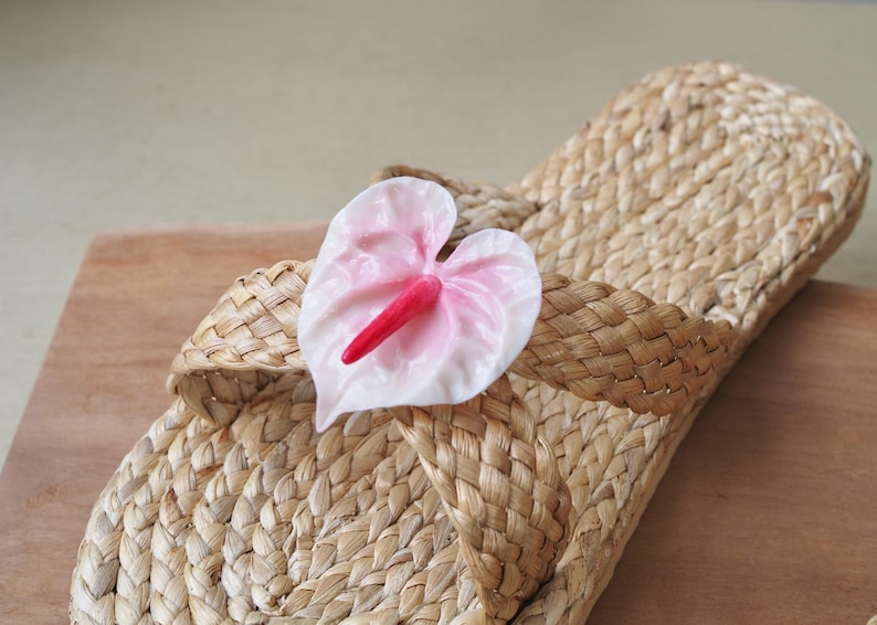 May include: A pair of woven straw sandals with a pink flower accent. The flower has a red center and is attached to the sandal with a woven strap.