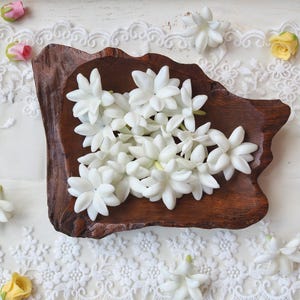 Puede incluir: Una colección de flores de jazmín blancas dispuestas dentro de un cuenco de madera de color marrón oscuro con forma irregular. El cuenco está rodeado de pequeños capullos de rosa amarillos y rosas y tela de encaje blanco con patrones florales.