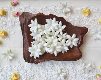 May include: A collection of white jasmine flowers arranged inside a dark brown, irregularly shaped wooden bowl. The bowl is surrounded by small yellow and pink rosebuds and white lace fabric with intricate floral patterns on a white surface.