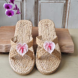 May include: A pair of woven straw sandals with pink flower accents. The sandals have a criss-cross strap design and a flat sole.