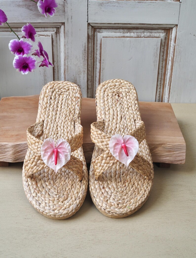 May include: A pair of woven straw sandals with pink flower accents. The sandals have a criss-cross strap design and a flat sole.