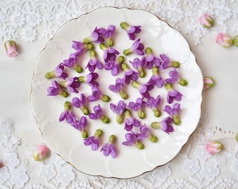 May include: A collection of small, purple lilac-shaped floral embellishments with green stems, scattered across a white scalloped plate. The flowers feature four distinct petals and a compact, rounded base, showcasing a vibrant violet hue.