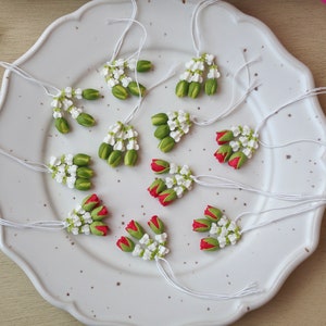 May include: A white plate with ten handmade flower garlands. The garlands are made of white and green polymer clay and have red accents. The garlands are strung on white thread.