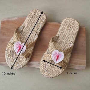 May include: Two pairs of woven straw sandals with pink flower embellishments. The sandals are 10 inches long and 3 inches wide.