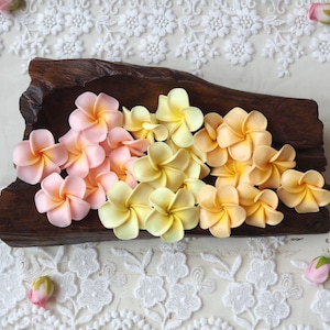 Puede incluir: Una colección de flores de plumeria artificiales en tonos rosa suave, amarillo pálido y melocotón cálido, dispuestas dentro de un cuenco de madera de color marrón oscuro y aspecto rústico. Las flores tienen cinco pétalos redondeados.