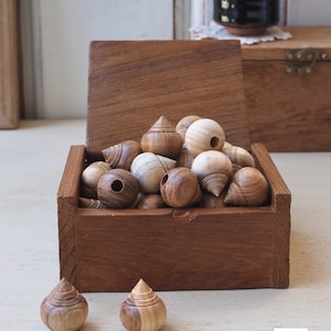 Pode incluir: Uma caixa de madeira cheia de contas de madeira de diferentes formas, algumas redondas e outras com forma de lágrima. Duas contas adicionais estão fora da caixa sobre uma superfície de madeira clara.