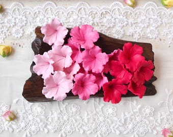 May include: A collection of artificial flowers in varying shades of pink, ranging from light pastel to deep magenta, arranged on a dark brown piece of wood. The flowers feature five rounded petals each and are displayed in a gradient color pattern.
