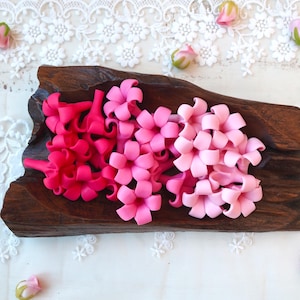 Puede incluir: Una bandeja de madera contiene grupos de flores artificiales rosas y fucsias. Las flores están dispuestas en un degradado de tonos, desde fucsia intenso hasta rosa claro. La bandeja se encuentra sobre una superficie de encaje blanco, con capullos florales esparcidos.