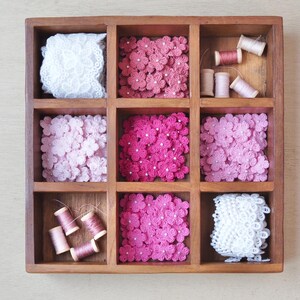 May include: A wooden box with nine compartments, each filled with pink and white crocheted flowers and spools of thread. The box is a great way to organize and store craft supplies.