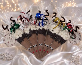 May include: Five clear plastic cone-shaped bags filled with hot cocoa mix, dark chocolate chips, and white mini marshmallows. Each bag features googly eyes, a red nose, brown pipe cleaner antlers, and colorful curling ribbons at the top.