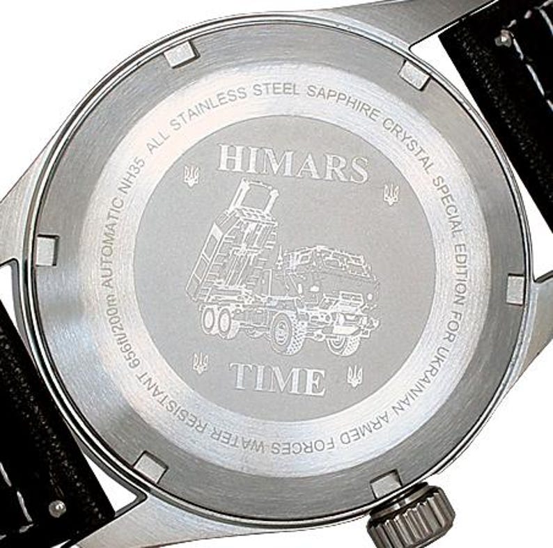 Puede incluir: La parte posterior de un reloj plateado con una correa de cuero negro. La esfera del reloj tiene las palabras "HIMARS" y "TIME" grabadas. El reloj es una edici&oacute;n especial para las Fuerzas Armadas de Ucrania.