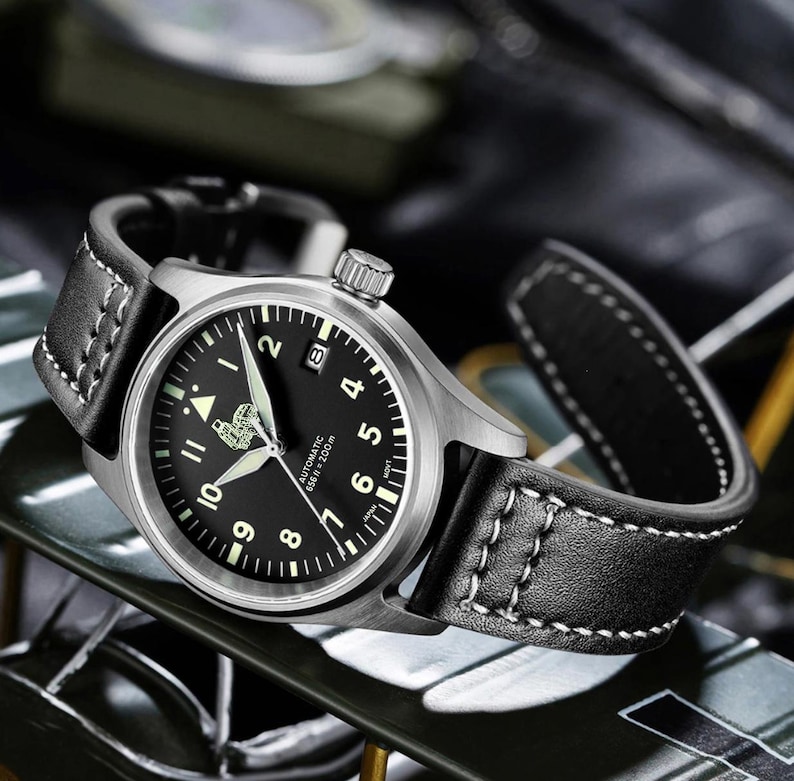 Puede incluir: Un reloj de pulsera de cuero negro con caja plateada y esfera negra. El reloj tiene marcas blancas y una ventana de fecha en la posici&oacute;n de las 3 en punto. La esfera del reloj tambi&eacute;n incluye el texto "Automatic 65ft = 200m".